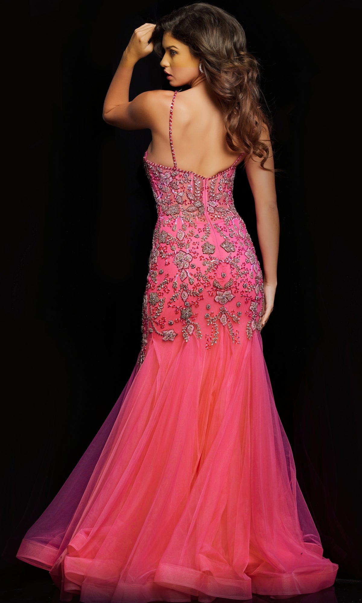 A woman faces away from the camera in the Jovani Beaded Sheer-Bodice Long Prom Dress 23125, a bright pink gown with floral embroidery and bead details, set against a black background.